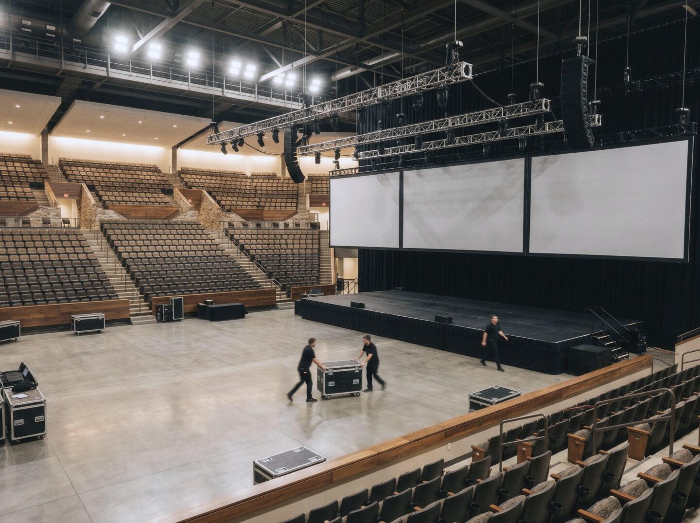 Kona Arena during setup for a general session, with crew pushing a road case, a wide stage, blank screens, and tiered seating.