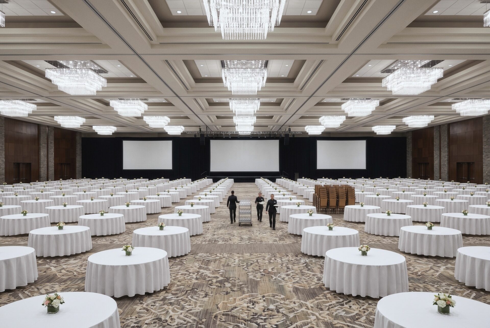 The Pacific Grand Ballroom set for a banquet, with staff walking an aisle among hundreds of round tables, a wide stage with screens, and a high coffered ceiling.