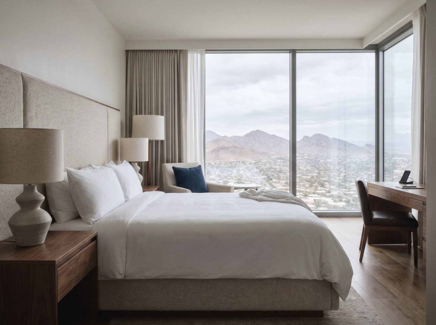 A Resort King room with a white king bed, a walnut nightstand and lamp, a desk, and a window onto desert mountains.