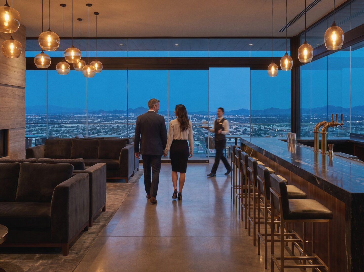Skyline Lounge at dusk, with a couple walking past the long stone bar toward a wide view over the city to the mountains.